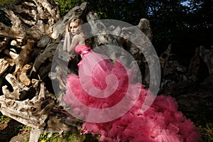 Charming woman pink evening dress with fluffy aerial skirt is posing in botanical garden on the driftwood dried wood trunks