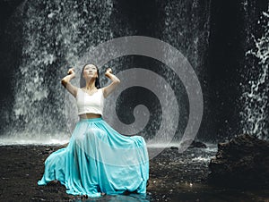 Charming woman sitting near waterfall. Water splashing. Travel lifestyle. Woman wearing long blue skirt. Nature in Bali