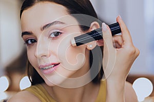 Charming woman using concealer stick while putting makeup on