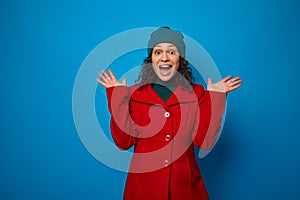Charming wonderful pretty woman in bright red coat and green hat looking surprised at the camera and holding hands palms up,