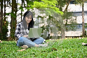 Charming young Asian female college student using laptop in the campus park