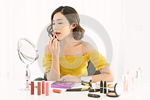 Charming young Asian woman wearing off shoulder yellow knitted clothes sit in front of mirror and applying red lip tint with her