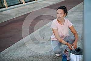 Charming young athlete sits next to a bike path and a treadmill on a bridge, pulling her sports equipment from her sports backpack