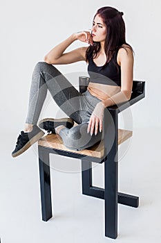 Charming young athletic sportwoman in sportsuit is posing and looking aside while sitting on the chair in white studio