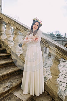 Charming young bride in long white wedding dress and floral wreath standing back on the old stone stairs
