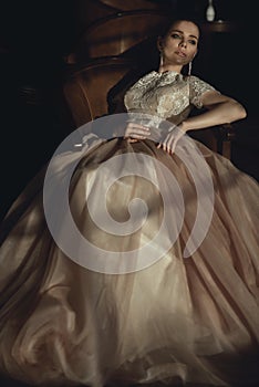 Charming young bride in luxurious designer dress with pink veiling skirt sitting relaxed in the old brown armchair