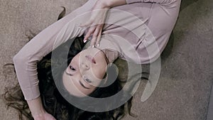 Charming young brunette woman is lying on carpet in room, top view portrait in apartment, looking at camera