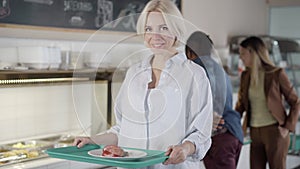 Charming young Caucasian blond woman holding tray with healthful vegetarian food looking at camera, and leaving