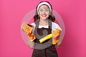 Charming young Caucasian housewife wearing headband, rubber gloves and apron, holding sponge and detergent in hands, looks smiling