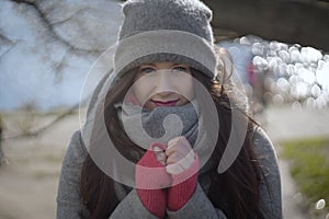 Charming young Caucasian woman holding hands together on sunny autumn day. Portrait of brunette girl in warm clothes