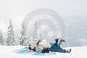Charming young couple in warm winter knitted sweaters lying on the wool blue chekered blanket in snowy forest. Love in winter, Hap