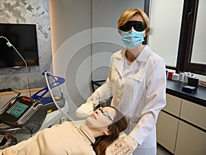 Charming young European woman getting facial laser treatment by cosmetician in wellness spa clinic