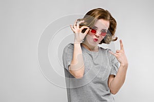 Charming young girl in a gray T-shirt on a gray background. Girl in red square-eyed glasses. The girl shows a rocker