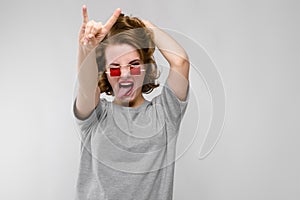 Charming young girl in a gray T-shirt on a gray background. Girl in red square-eyed glasses. The girl shows a rocker