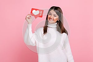 Charming young girl holding heart like icon of social media and looking at camera with toothy smile.