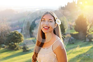 Charming young girl with shy smiling. Photo of beautiful young woman with flower on ear and natural background