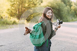 A charming young girl walks in nature, holding a backpack on her shoulder, from which her dog looks out, and holding a camera in