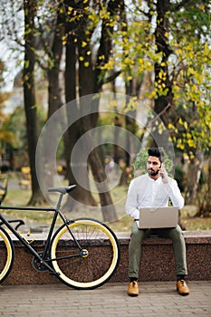 Charming young indian businessman talking on mobile phone while working on laptop computer sitting outdoors with bicycle