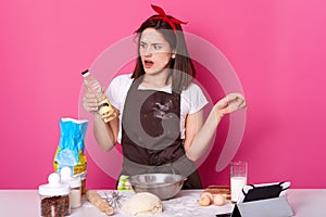 Charming young lady making bakery in kitchen, uses many ingredients and utensils for kneading dough, looks with astonishmant on