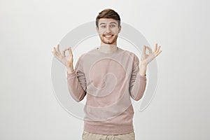 Charming young man with bright smile and bristle, wearing casual pullover and showing okay or zen gesture while standing