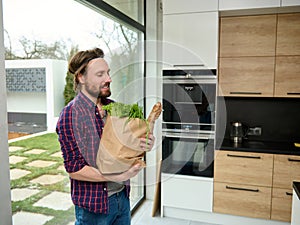 Charming young man enters the kitchen holding paper craft eco bag with food. Handsome guy with cardboard sack full of healthy food