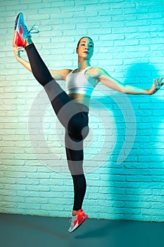 Charming young slim gymnast woman stretching on brick wall in neon lights. Flexible gymnastic exercise.