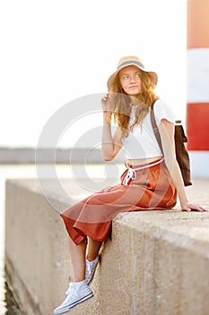 Charming young tourist woman sitting on background of lighthouse. Attractive red-haired girl traveling by Mediterranean coast.