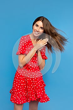 Charming young woman with bright smile in red dress feeling happy over blue background.