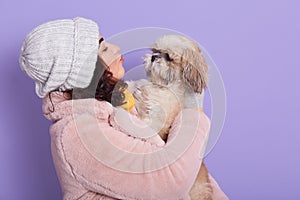 Charming young woman with dark hair, holds favorite dog, plays with Pekingese, dressed in faux fur coat, isolated over lilac