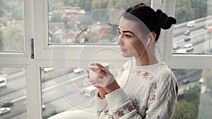 Charming young woman in earphones sitting near window listening musik and drink coffee