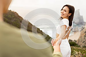 Charming young woman holding man& x27;s hand, enjoying romantic date on coastline over ocean beach