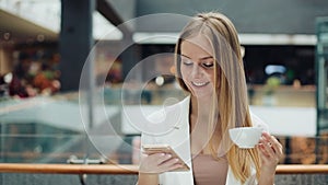 Charming young woman holds smartphone in one hand and cup of coffee in another sitting in the cafe