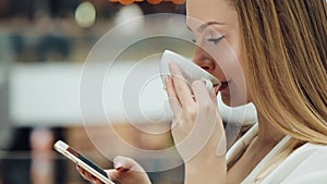 Charming young woman holds smartphone in one hand and cup of coffee in another sitting in the cafe