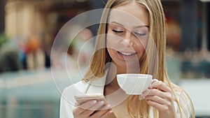 Charming young woman holds smartphone in one hand and cup of coffee in another sitting in the cafe