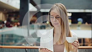 Charming young woman holds smartphone in one hand and cup of coffee in another sitting in the cafe