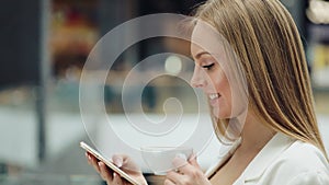 Charming young woman holds smartphone in one hand and cup of coffee in another sitting in the cafe