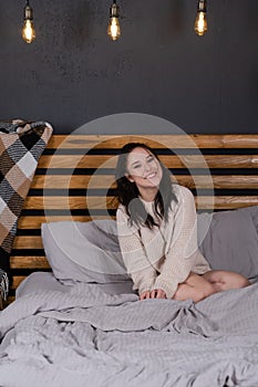 charming young woman in knitted beige sweater in bed with grey linens. modern loft bedroom in brown and gray colors