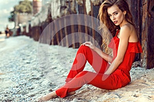 Charming young woman with long hair in coral red one shoulder jumpsuit sitting on the beach at the old rusty piles