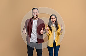 Charming young woman and man dressed in casuals showing thumbs up and laughing while standing over beige background. Cheerful