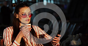 Charming young woman in red sunglasses drinks coffee in the rays of evening sun