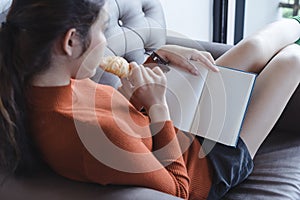 Charming young woman sitting on sofa eating a croissant and reading a book. blank book for graphic montage.