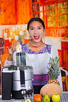 Charming young woman in traditional andean dress standing inside kitchen inserting apple pieces into juice maker