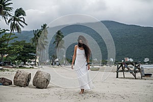 Charming young woman in white dress walking on the beach and looking down.