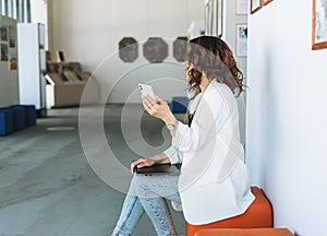 Charming young woman in white suit using mobile with laptop in the modern art gallery