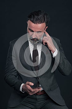 Chat with client. Good looking young man in full suit using his smart phone and smiling while standing against grey