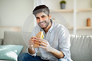 Cheat meal day. Happy arab man eating tasty sandwich and smiling to camera, sitting on sofa in living room at home