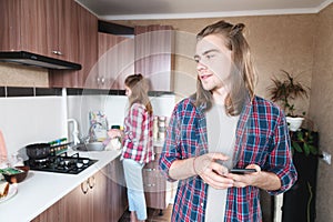 Cheating wife. The young man looks suspiciously at his wife who washes the dishes. The man has his smartphone in his