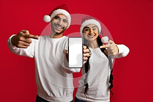 Check This App. Arab Couple Wearing Santa Hats Pointing At Blank Smartphone