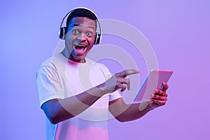 Check This App. Excited Black Man In Headphones Pointing At Digital Tablet