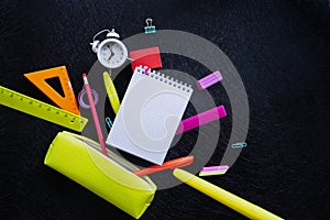 Checkered notepad, alarm clock and multicolored stationery falling out of a bright yellow pencil case against a black  background